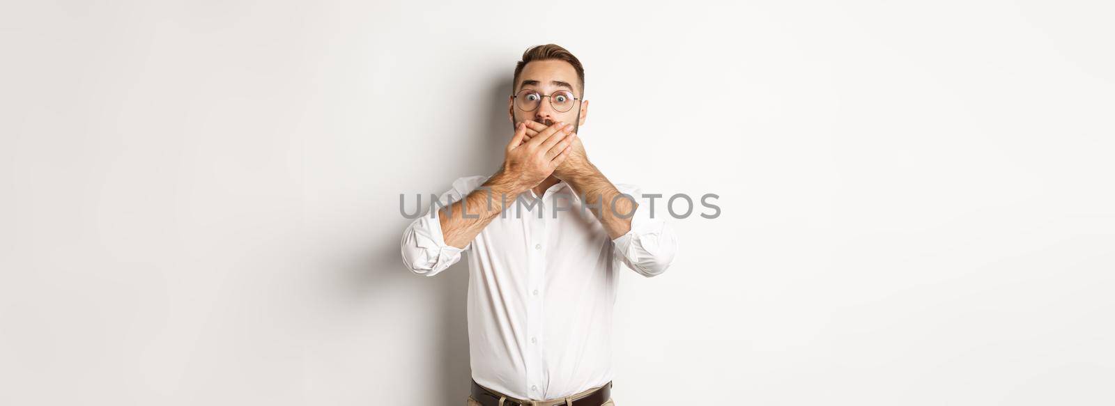 Shocked man gasping and looking at something in awe, covering mouth with hands, white background.