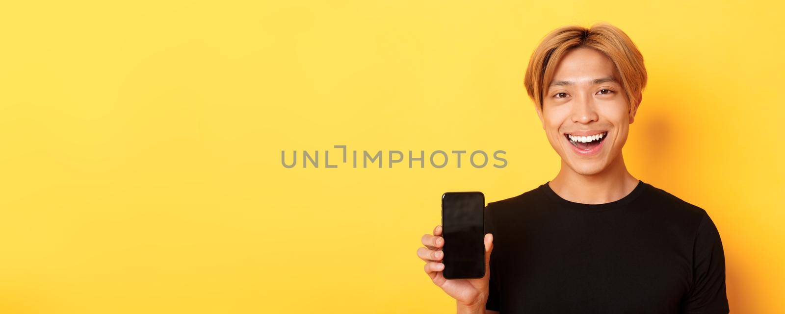 Close-up of stylish handsome korean guy showing smartphone screen and smiling pleased, recommend mobile app, standing over yellow background.