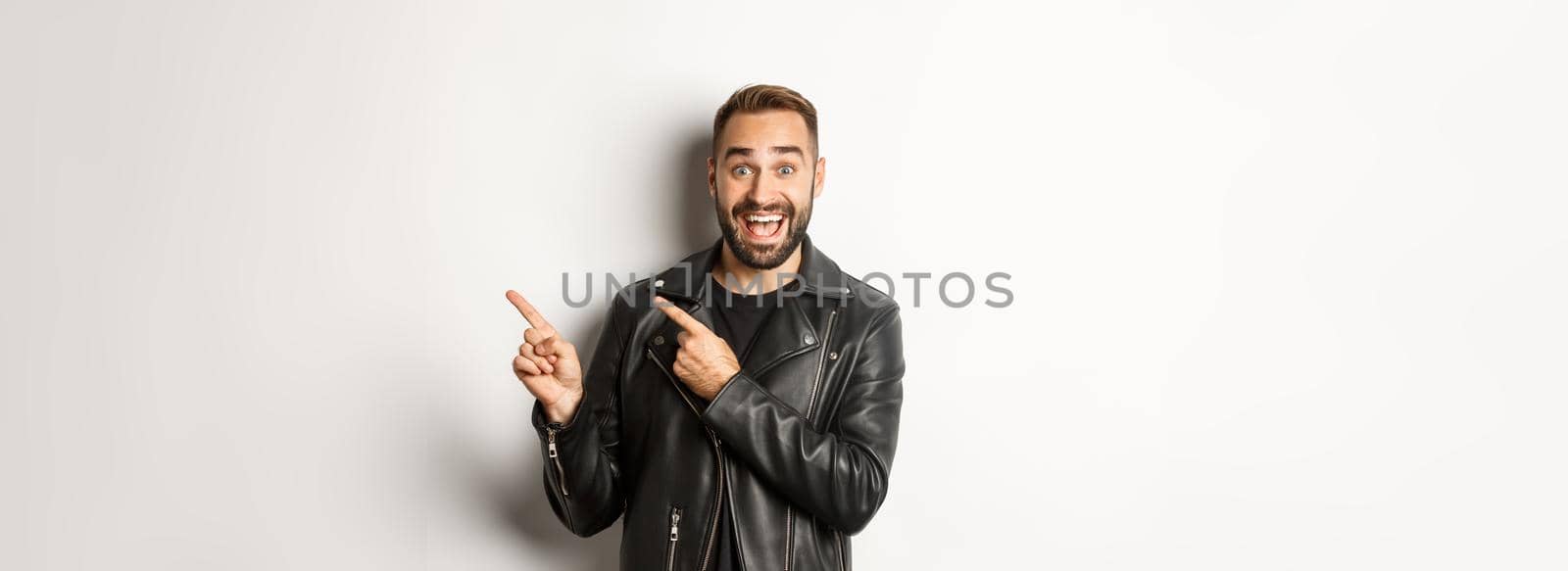 Excited, handsome man in black leather jacket, pointing fingers right at promo offer, showing logo, white background.
