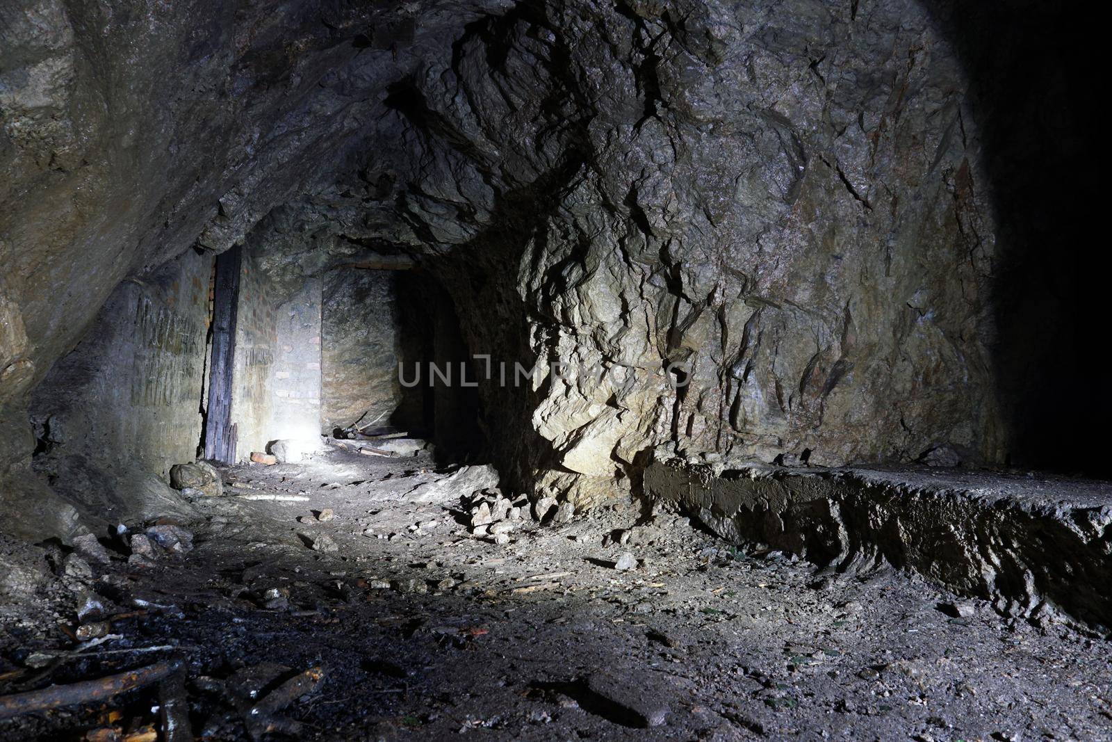 A dark artificial stone cave in the mountains. The light from the lantern falls on the rocks, where there are passages in the form of doors. Everything is wet. There are old branches and bricks.