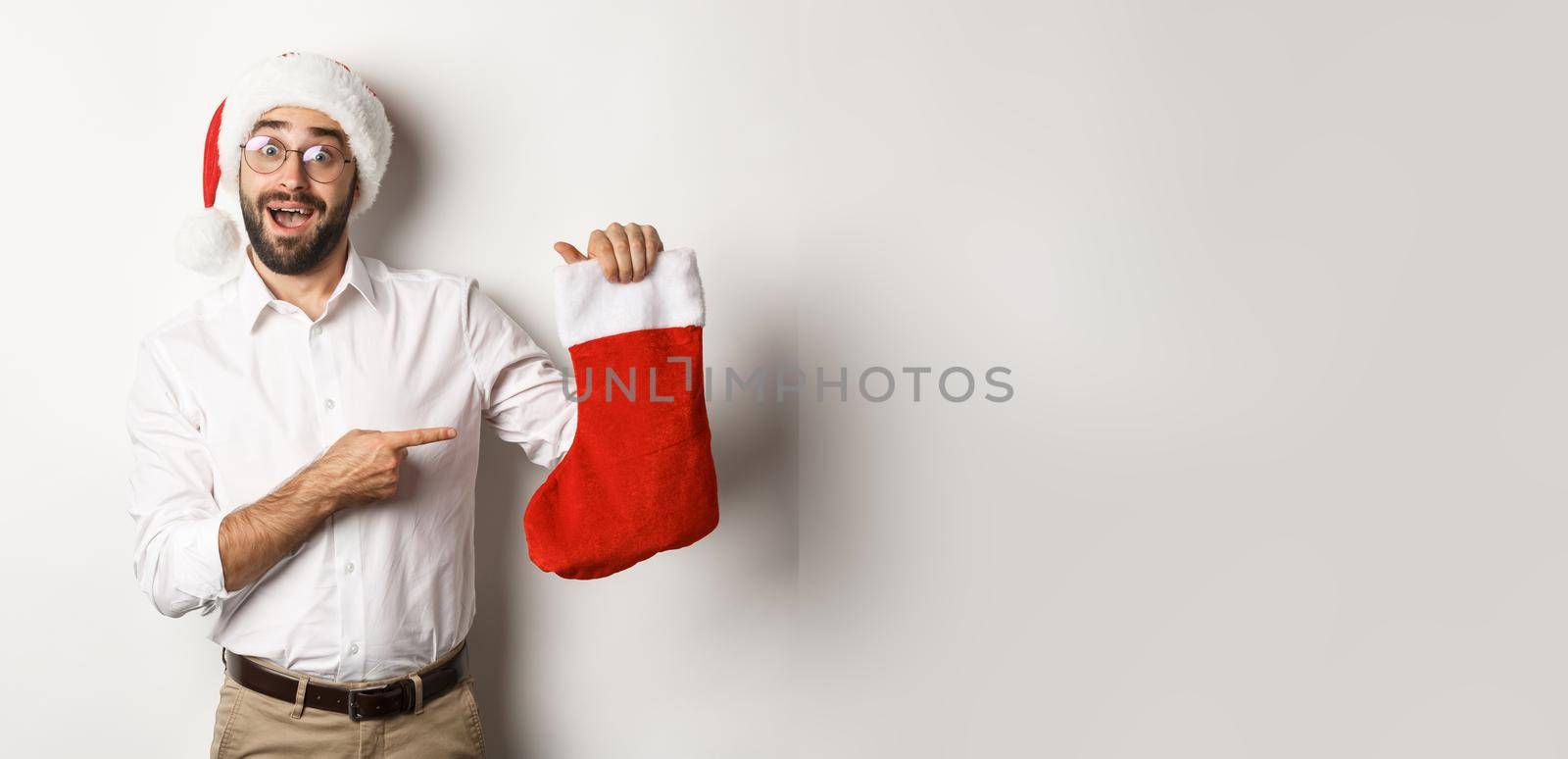Merry christmas, holidays concept. Happy adult man receive gifts in xmas sock, looking excited, wearing santa hat, white background by Benzoix