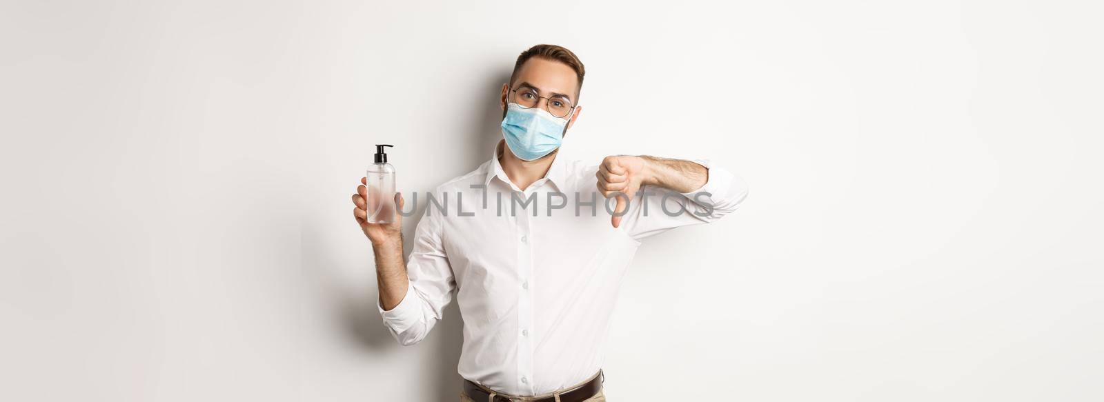 Covid-19, social distancing and quarantine concept. Office worker in medical mask displeased, showing hand sanitizer and thumb down, standing over white background.
