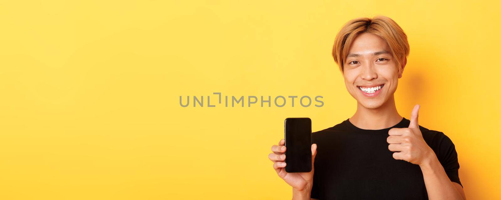 Satisfied handsomea young asian guy with fair hair, showing thumbs-up in approval and smartphone screen, standing over yellow background.