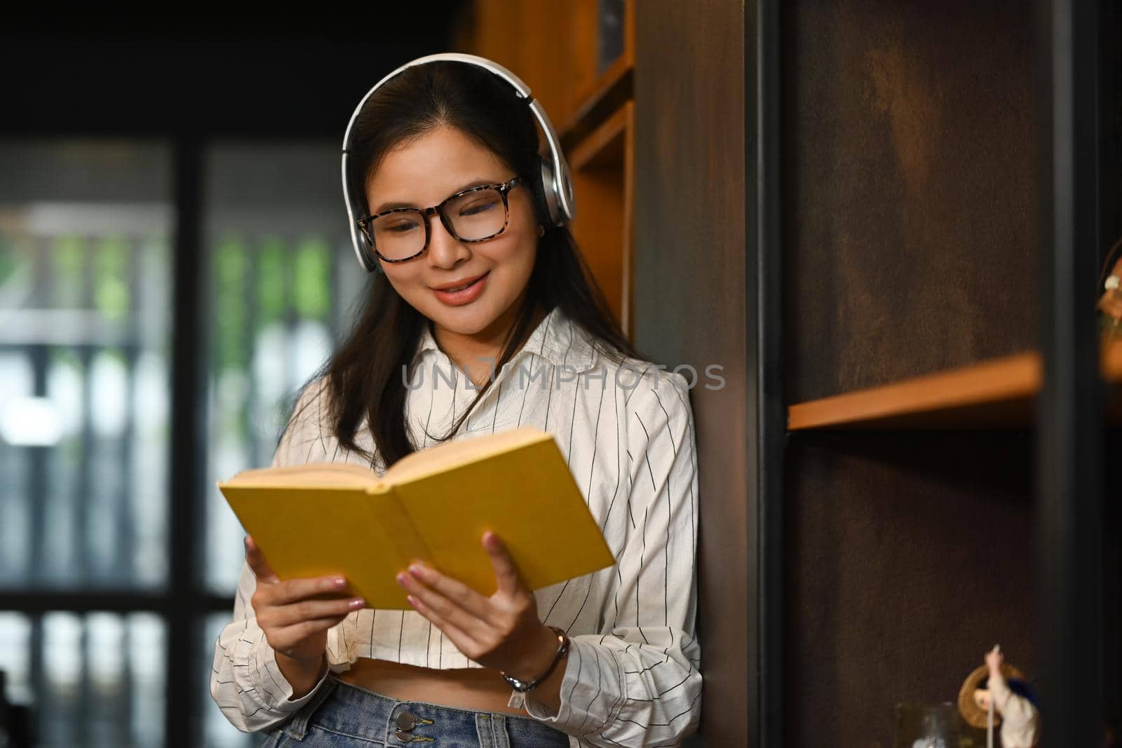 Portrait of college woman wearing headphone and reading book in library. People, knowledge and education concept by prathanchorruangsak