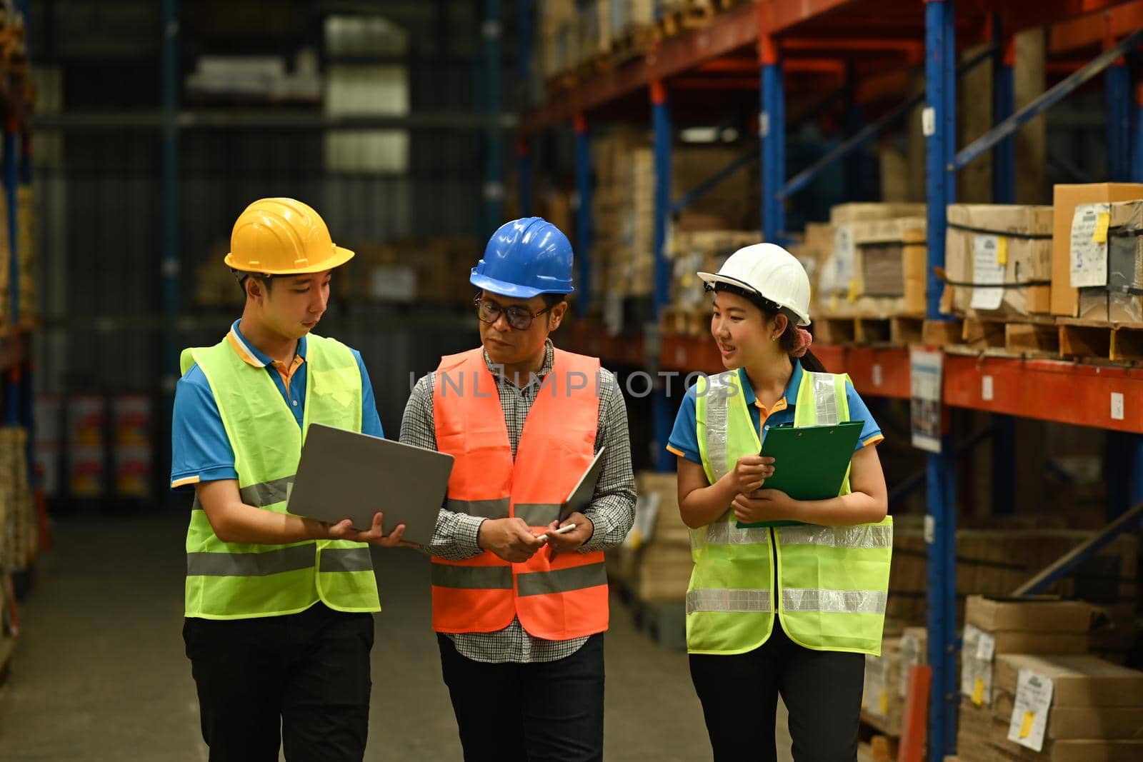 Middle aged manager and young workers inspecting stock and cardboard stock product on laptop computer in a large warehouse by prathanchorruangsak