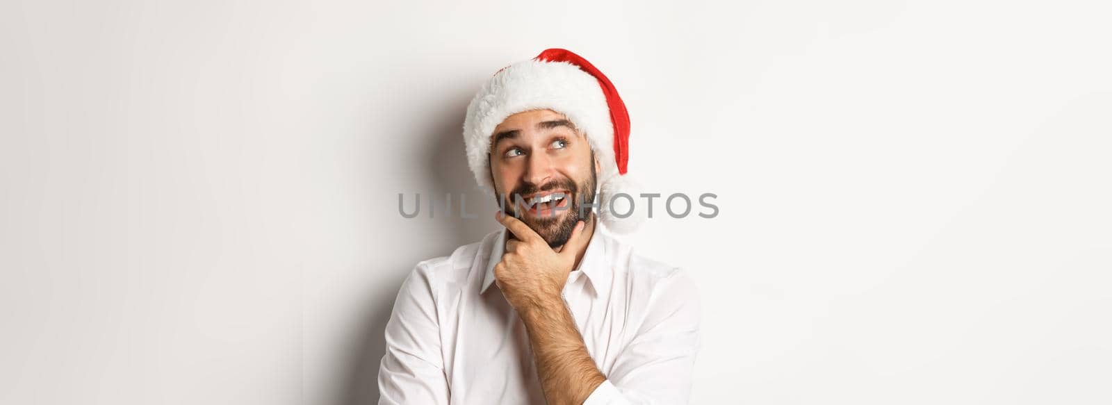 Party, winter holidays and celebration concept. Close-up of happy man planning christmas gift list, wearing santa hat, looking at upper left corner thoughtful, white background by Benzoix