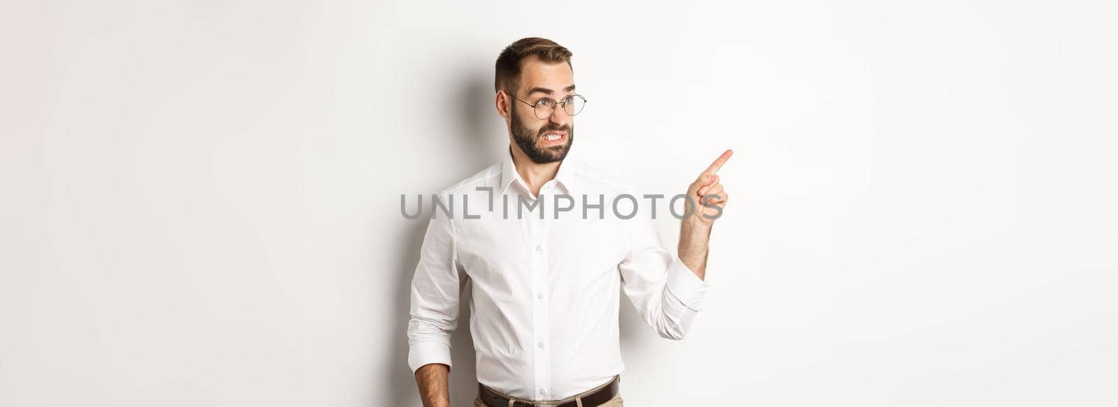 Young bearded guy seeing something disturbing, cringe while pointing finger left at promo offer, standing awkward against white background by Benzoix
