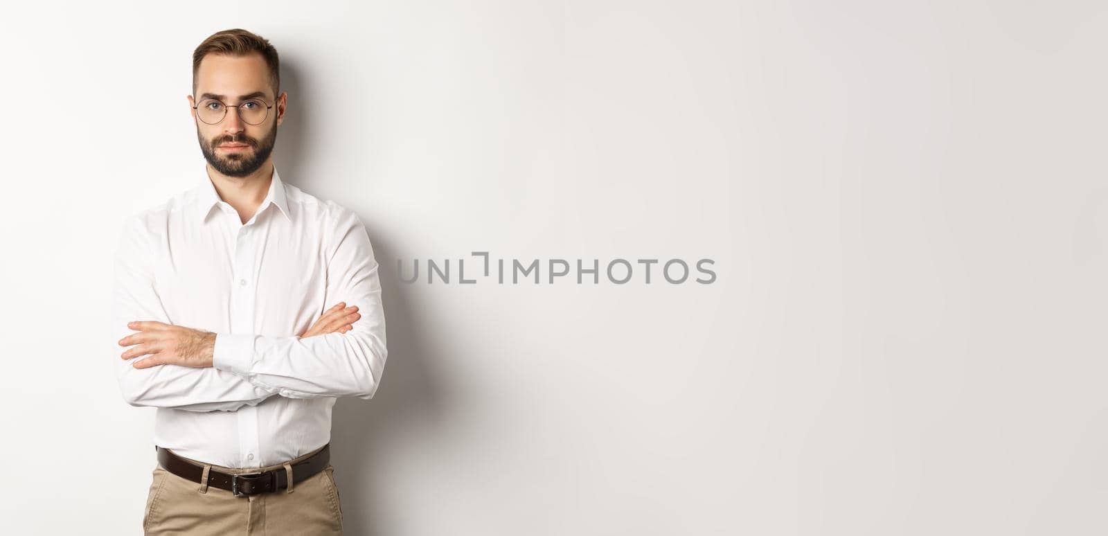 Confident businessman in glasses looking at camera, cross arms on chest, standing over white background.