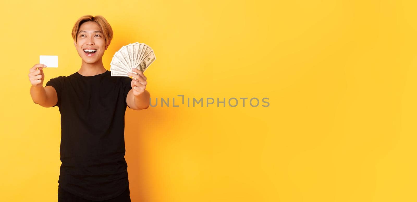 Happy good-looking asian guy looking thoughtful and pleased upper left corner while showing money and credit card, yellow background.