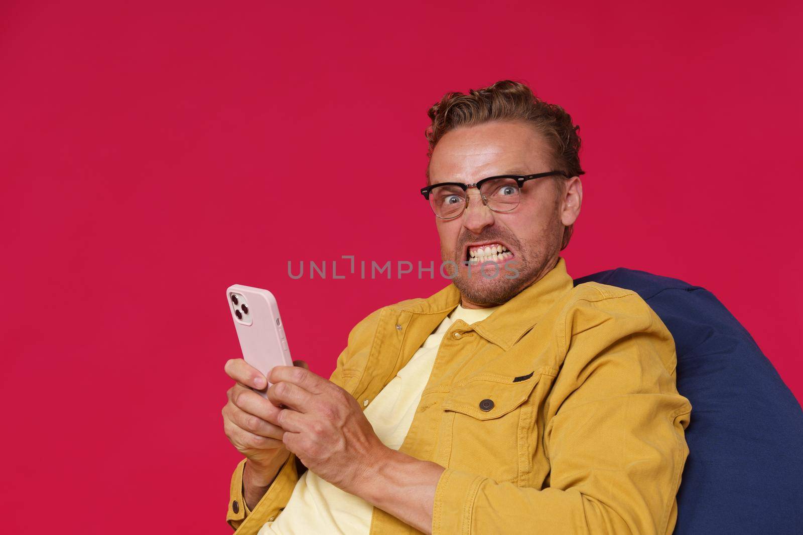 Not happy, annoyed handsome man in glasses hold smartphone in hands looking at camera with irritated look wearing yellow denim jacket sitting on bag chair isolated on red background.