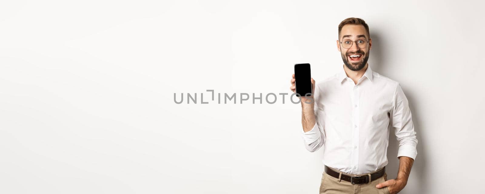 Amazed handsome businessman showing smartphone screen app, standing over white background.