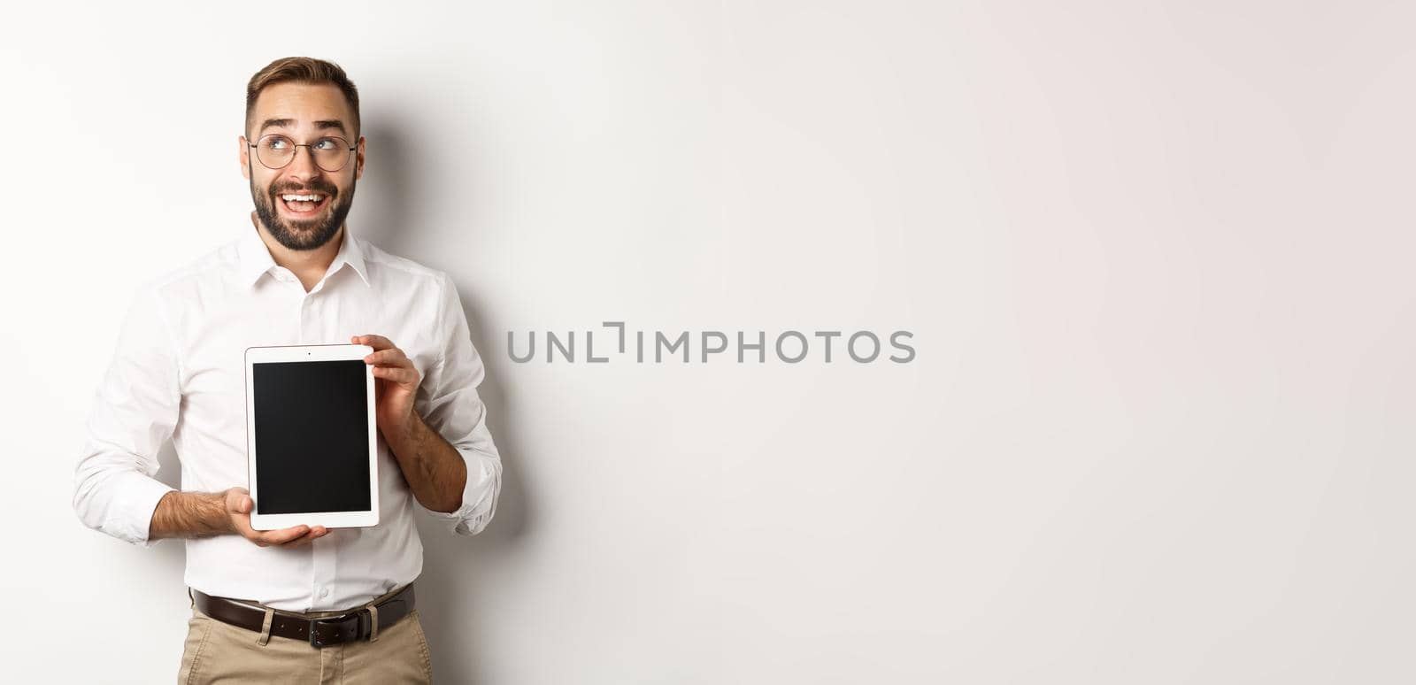 Shopping and technology. Thoughtful man showing digital tablet screen, looking at upper left corner and thinking, standing over white background.