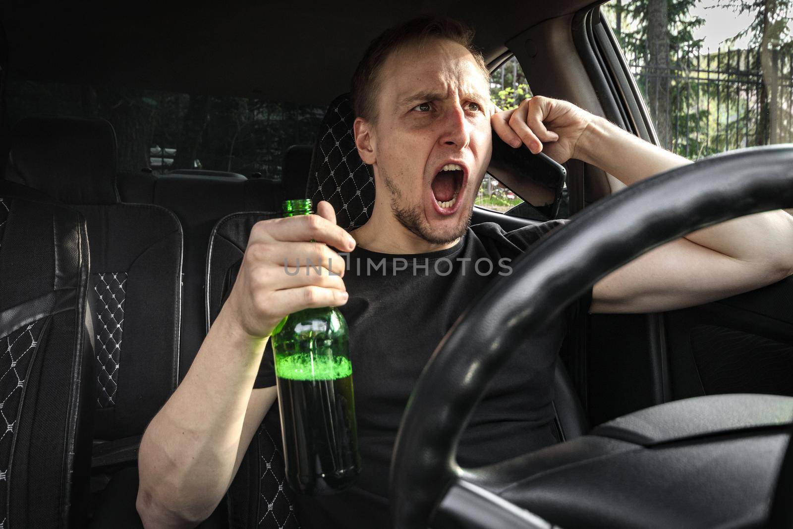 Emotional Drunk aggressive driver with a bottle of beer driving a car swears on the phone. Alcohol driving concept.