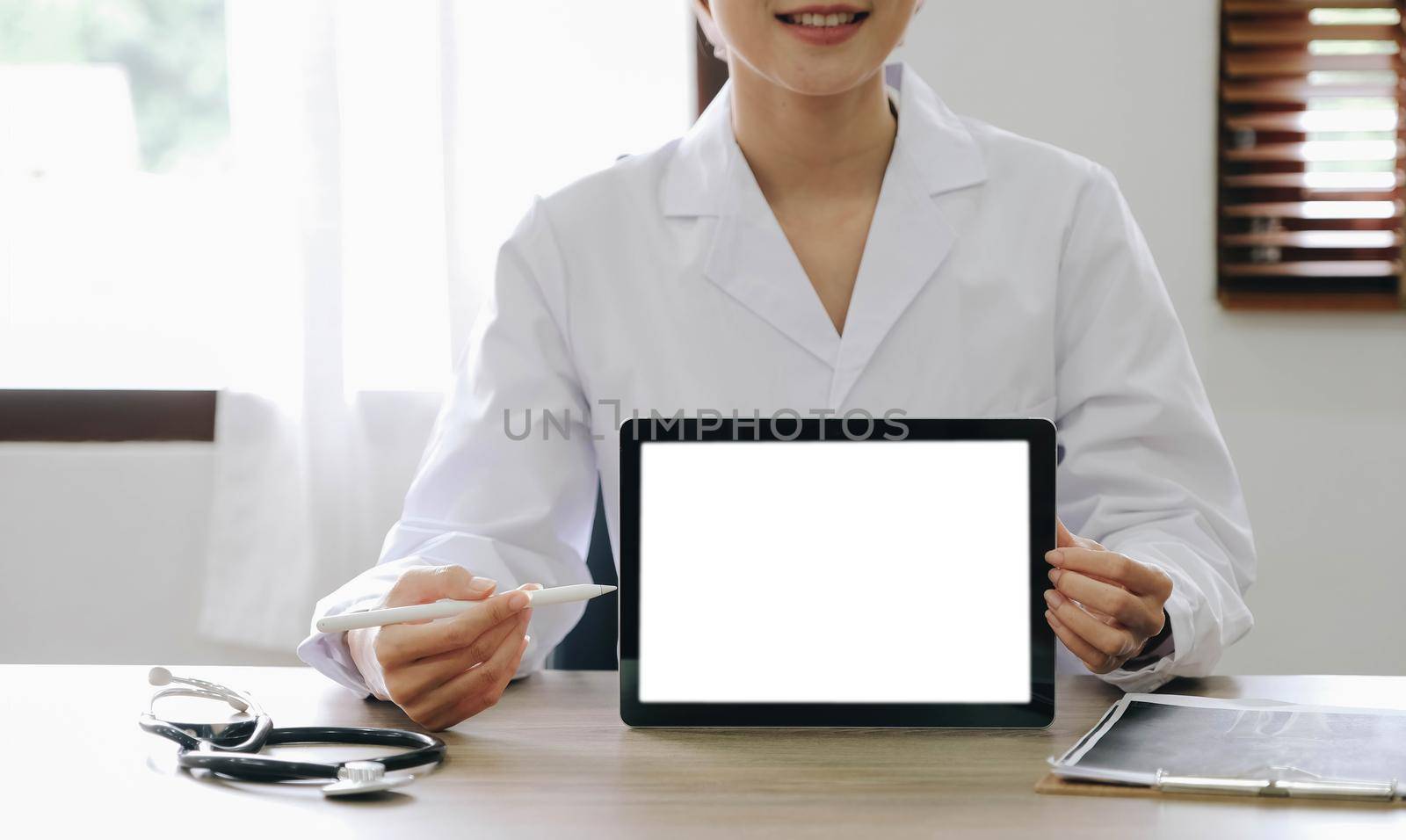 Portrait of experienced doctor woman cardiologist present modern technology white screen tablet she use consult patients wear white coat uniform stethoscope in examination room. by wichayada