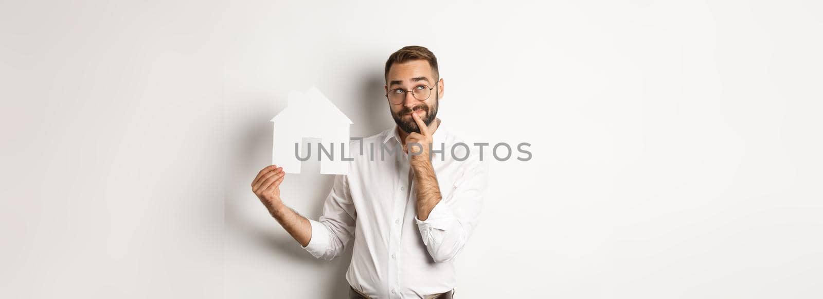 Real estate. Man thinking while searching for apartment, holding paper house model, standing over white background.