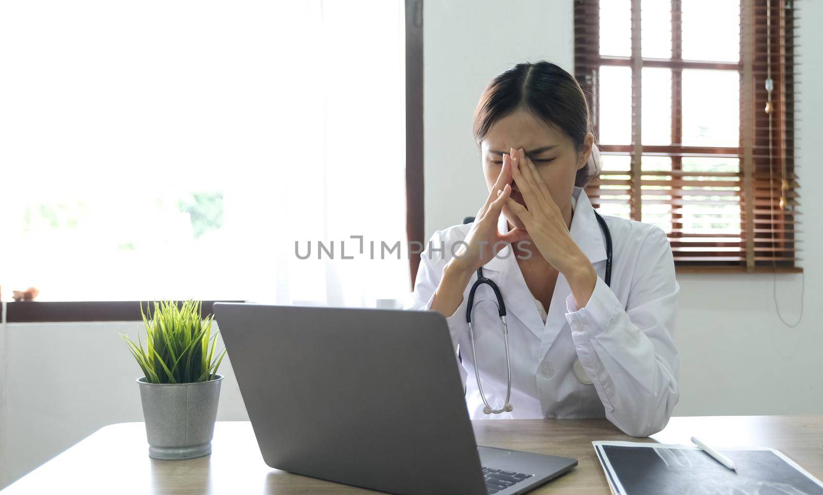 Female doctor holding stethoscope sitting with laptop stress headache about her job and work in hospital. by wichayada