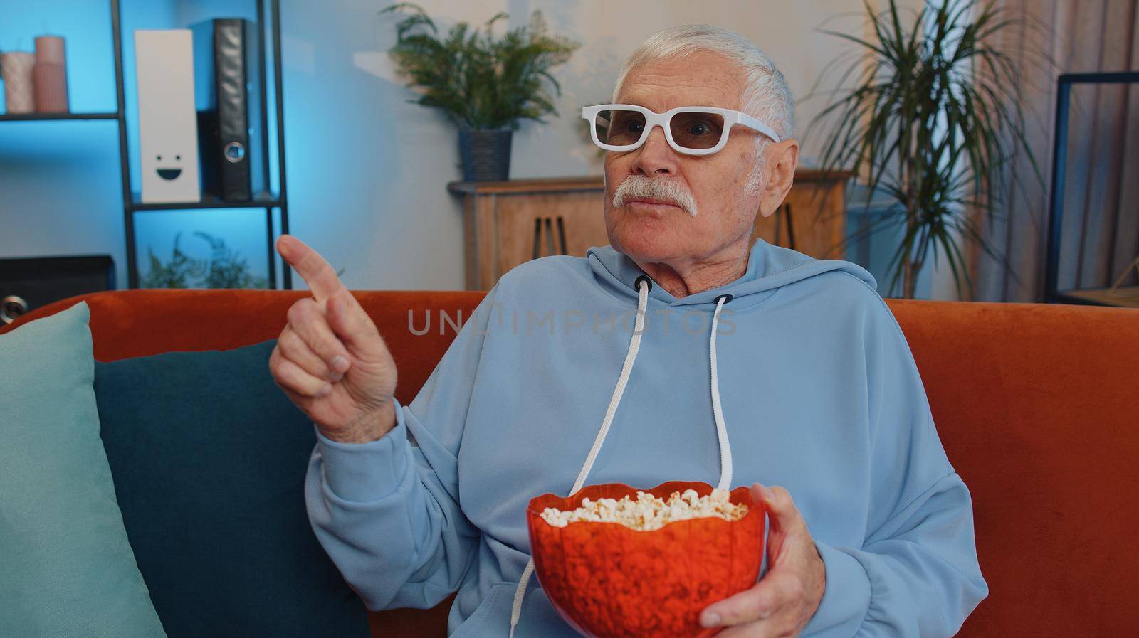 Excited elderly man on sofa eating popcorn and watching interesting tv serial, sport game, film, online social media movie content online at home. Senior grandfather enjoying domestic entertainment