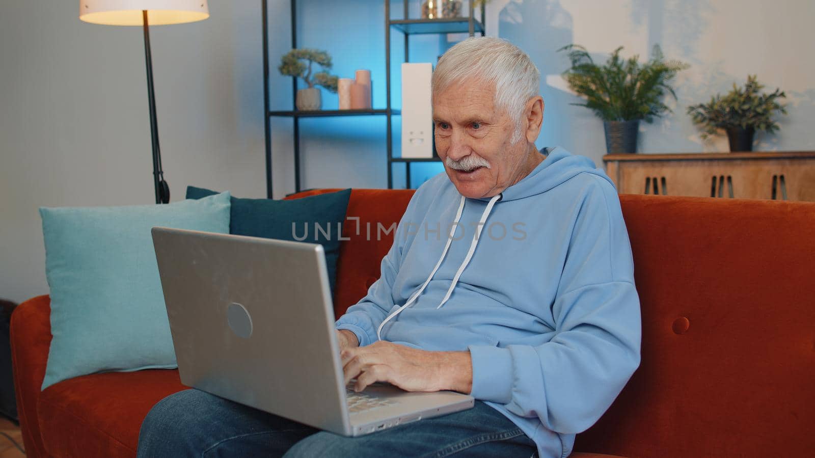 Mature elderly man freelancer at home living room sits on orange couch, opens laptop. Senior old retired grandfather works on notebook, sends messages, makes online purchases, watching movies, working