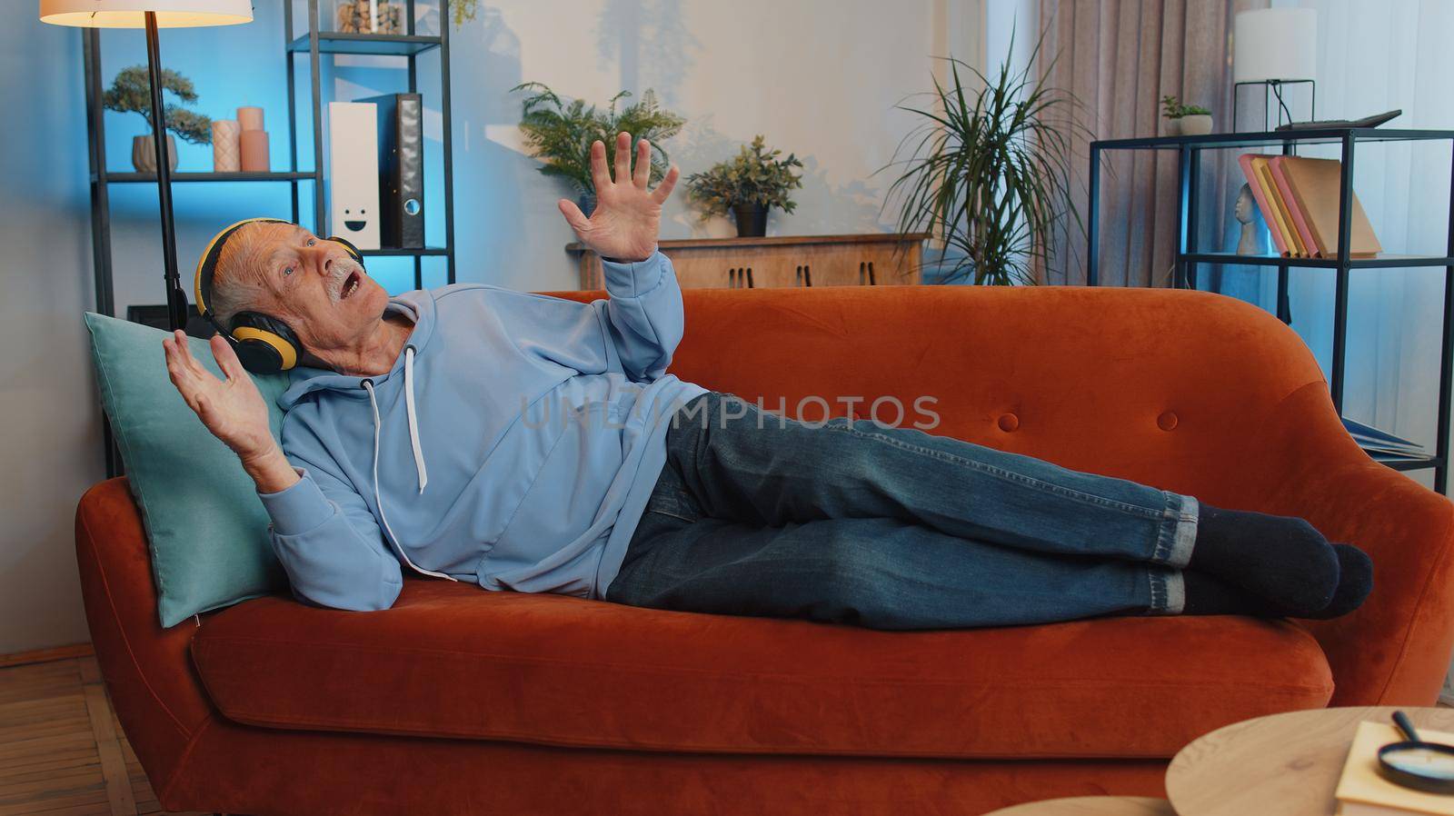 Senior old grandfather in wireless headphones relaxing lying on sofa at home choosing listening favorite energetic disco rock n roll music and dancing. People weekend activities. Happy overjoyed man