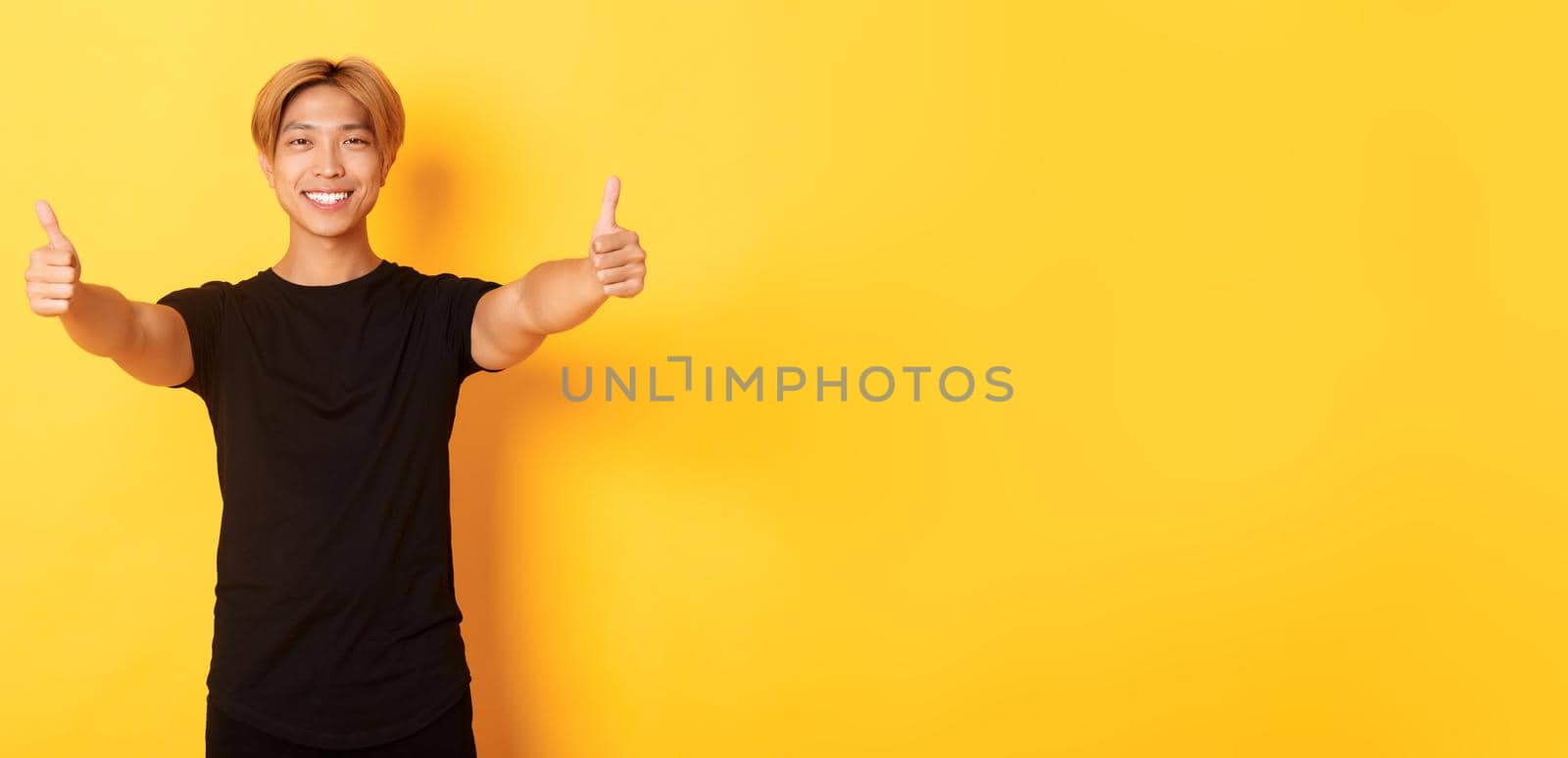 Portrait of assured handsome asian man showing thumbs-up in approval, like idea, standing yellow background by Benzoix