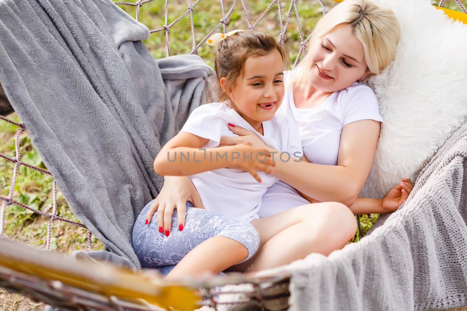 Beautiful young mother and daughter laying down and relaxing together on a hammock during a sunny summer day on holiday home garden. Family relaxing outdoors, healthy and wellness lifestyle.