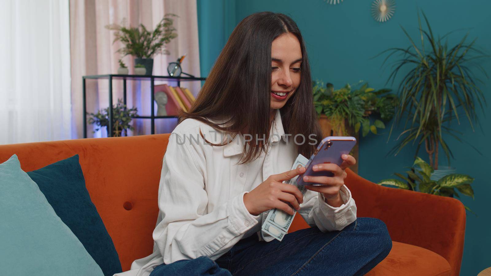 Planning family budget. Smiling caucasian girl counting money cash, use smartphone calculate domestic bills at home. Joyful young woman satisfied of income and saves money for planned vacation, gifts