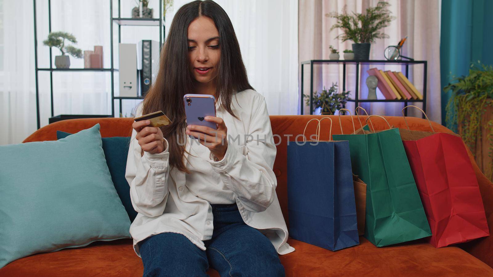 Happy shopaholic consumer girl sitting with shopping bags at modern home apartment making online order, payment with credit plastic bank card. Young woman on couch at home. Discount sale on internet