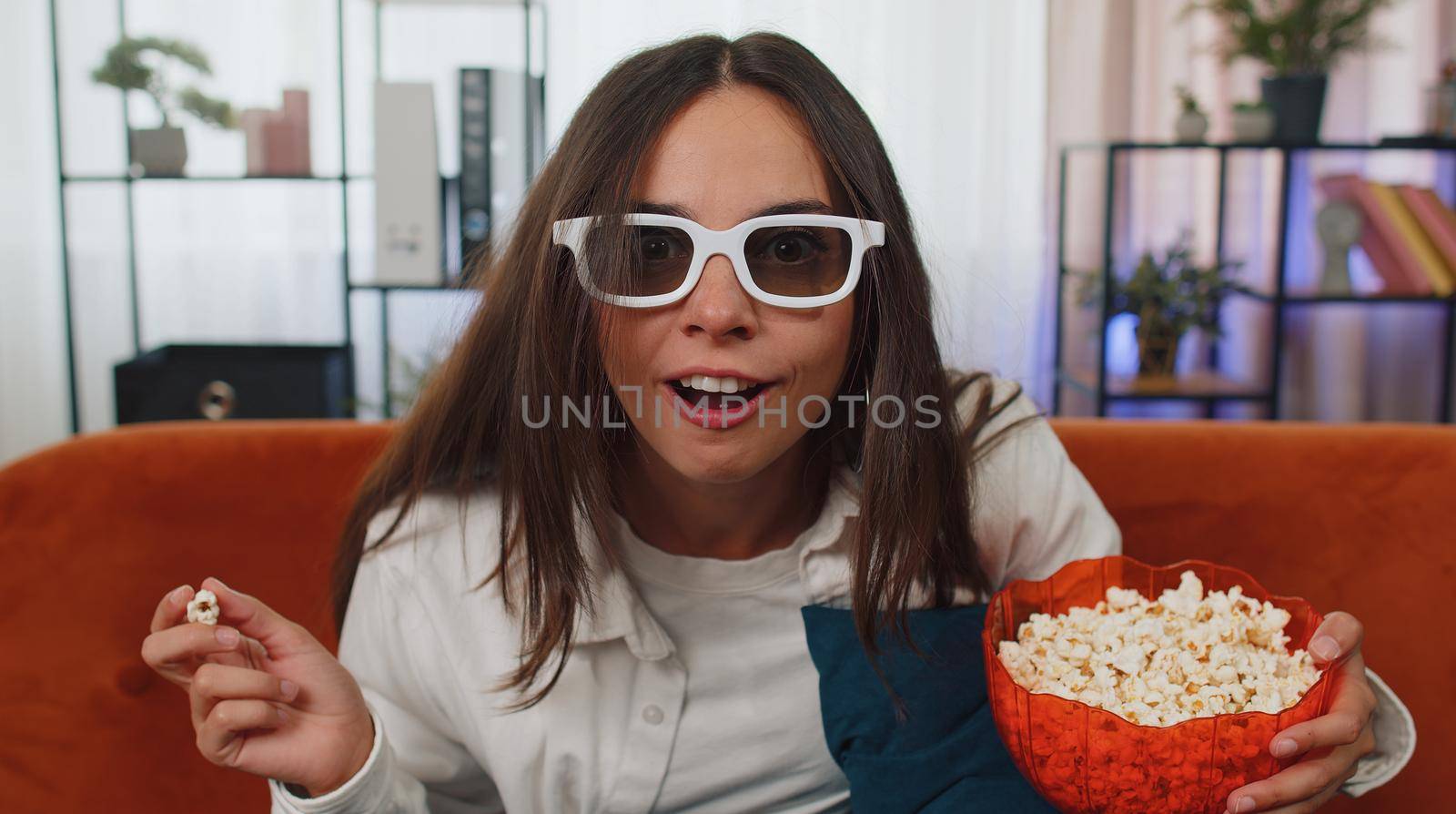 Excited adult girl sitting on sofa eating popcorn and watching interesting tv serial, sport game, film, online social media movie content online at home. Young woman enjoying domestic entertainment