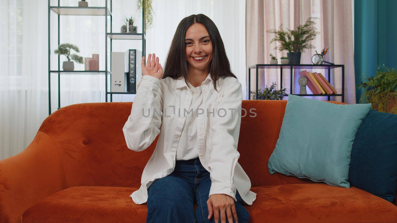 Portrait of caucasian adult girl smiling friendly at camera and waving hands gesturing hello, hi, greeting or goodbye, welcoming with hospitable expression. Young woman indoor at home in room on couch