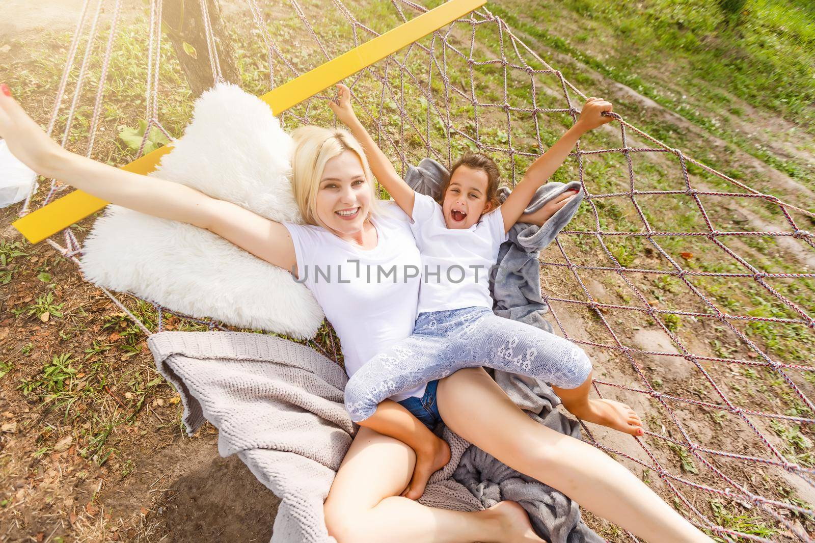 Beautiful young mother and daughter laying down and relaxing together on a hammock during a sunny summer day on holiday home garden. Family relaxing outdoors, healthy and wellness lifestyle.