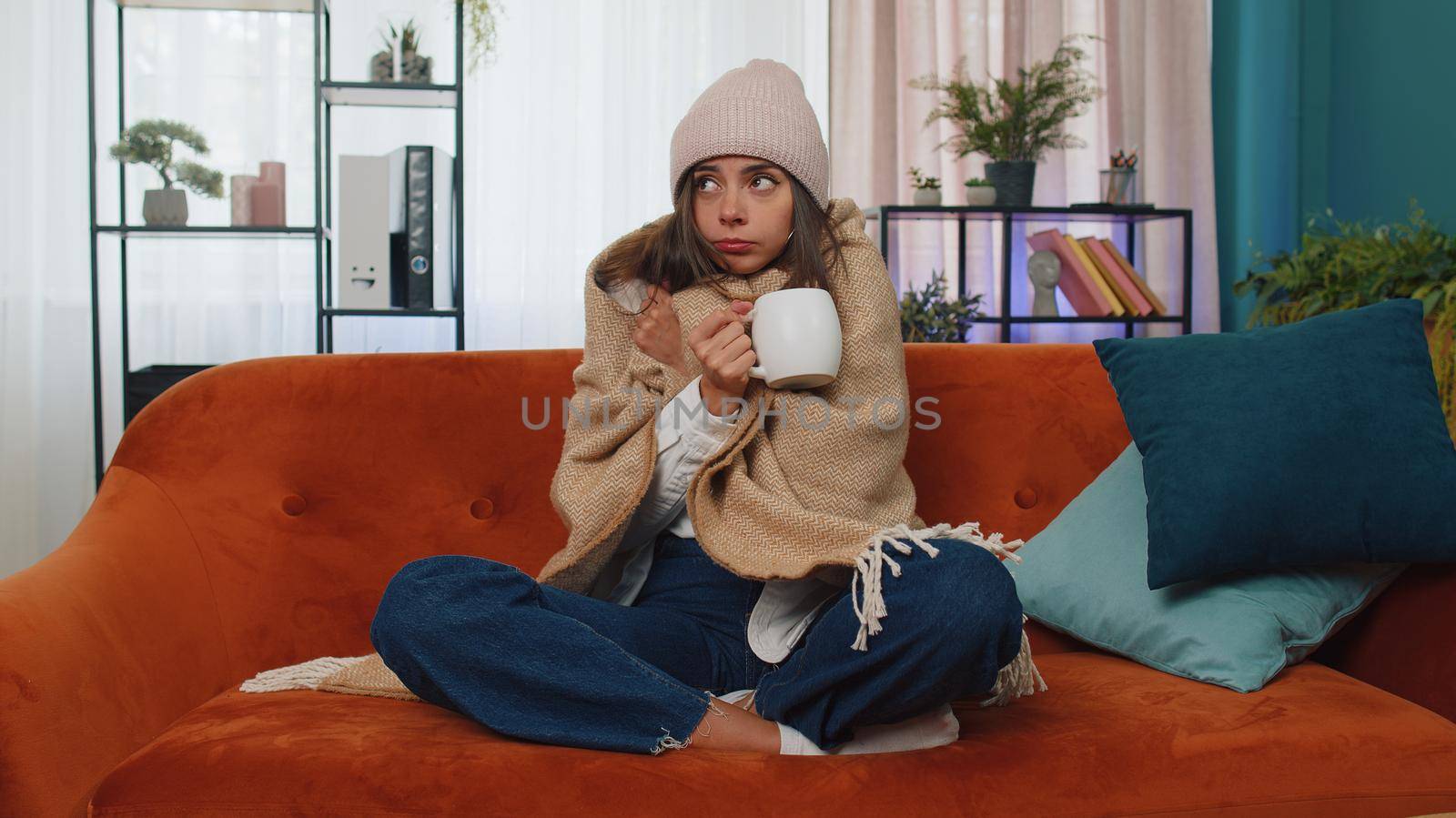 Sick girl wear hat wrapped in plaid sit alone shivering from cold on sofa drinking hot tea in unheated apartment without heating due debt. Unhealthy young woman feeling discomfort try to warming up