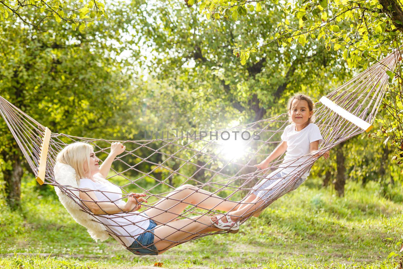 Happy mother and daughter relaxing together in a hammock at garden in sunny summer day. Family playing in hammock. by Andelov13