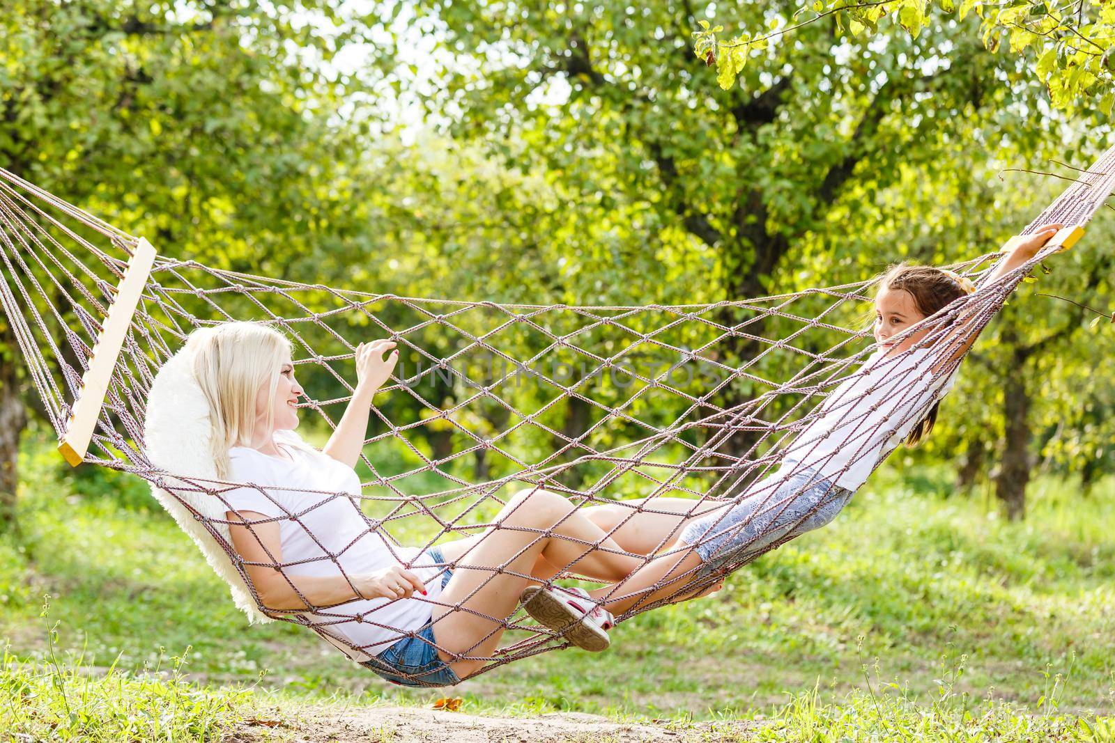 Happy mother and daughter relaxing together in a hammock at garden in sunny summer day. Family playing in hammock.