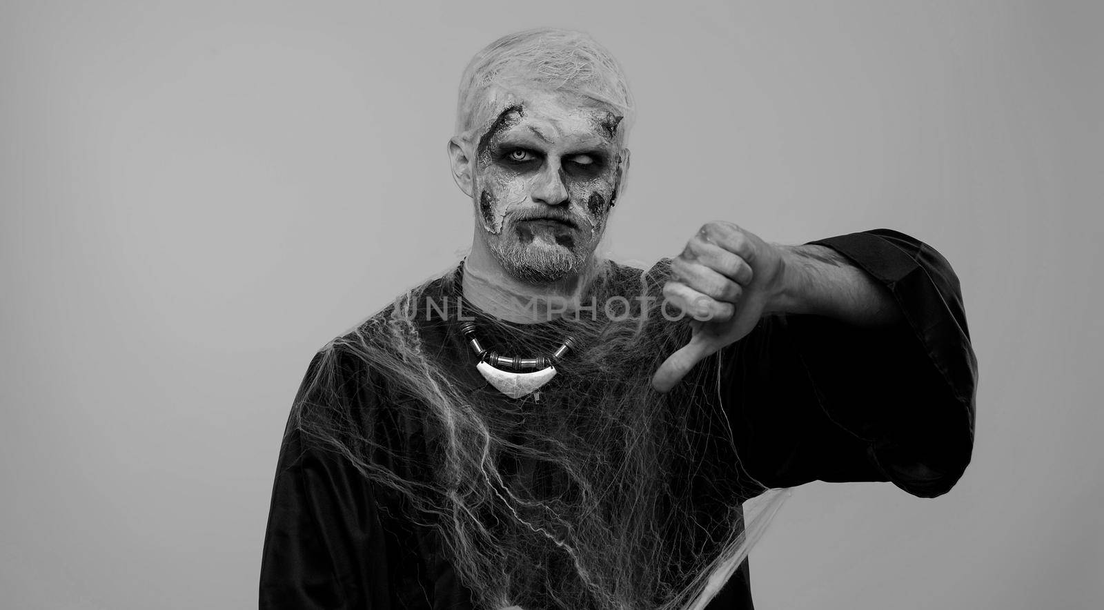 Sinister man in carnival costume of Halloween crazy zombie with bloody wounded scars face showing thumbs down sign gesture, expressing discontent, disapproval, dissatisfied, dislike. Cosplay undead