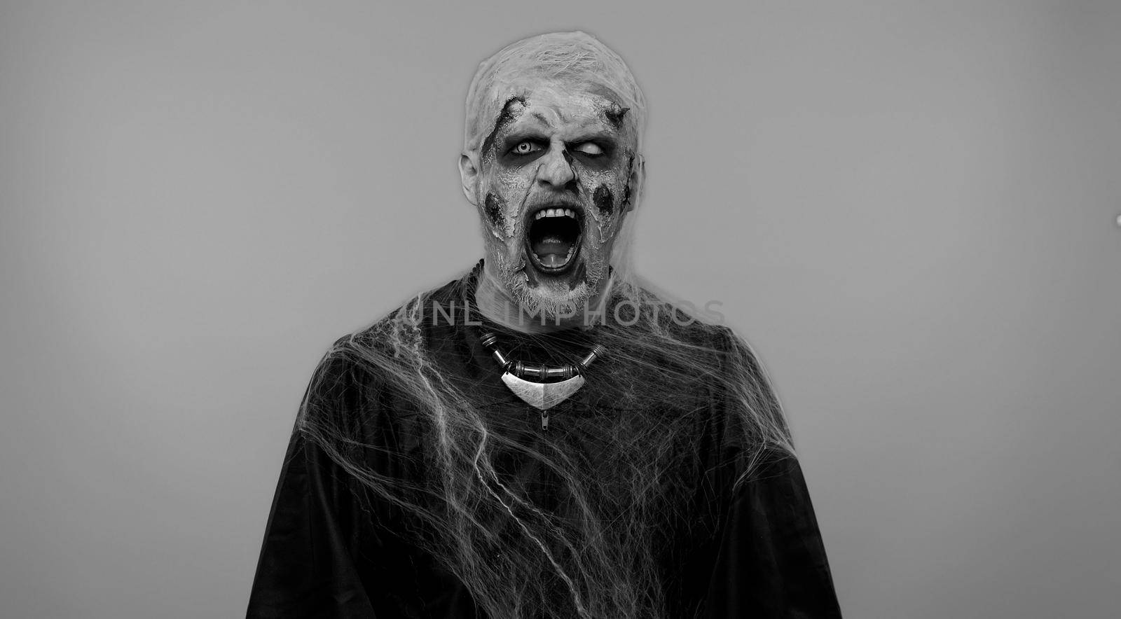 Sinister man in carnival costume of Halloween crazy zombie with bloody wounded scars face screaming, shouting, trying to scare on gray. Horror theme of cosplay wounded undead, beast, monster