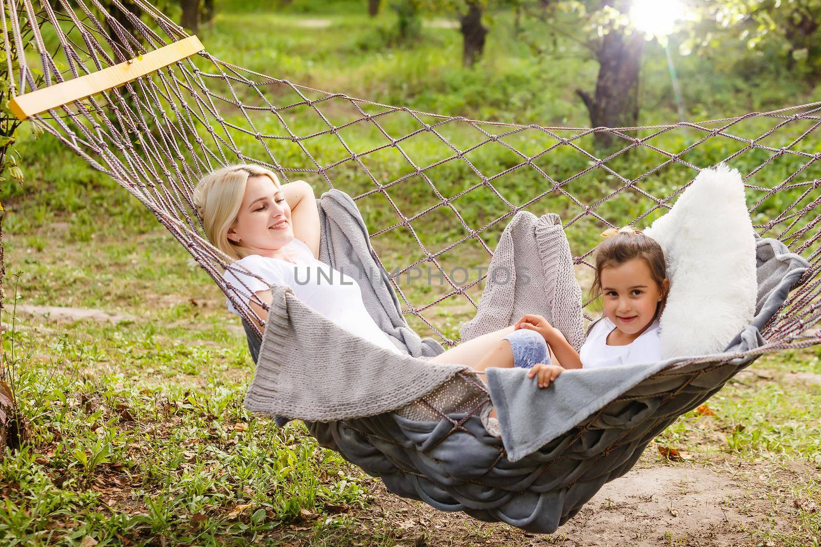 Beautiful young mother and daughter laying down and relaxing together on a hammock during a sunny summer day on holiday home garden. Family relaxing outdoors, healthy and wellness lifestyle.