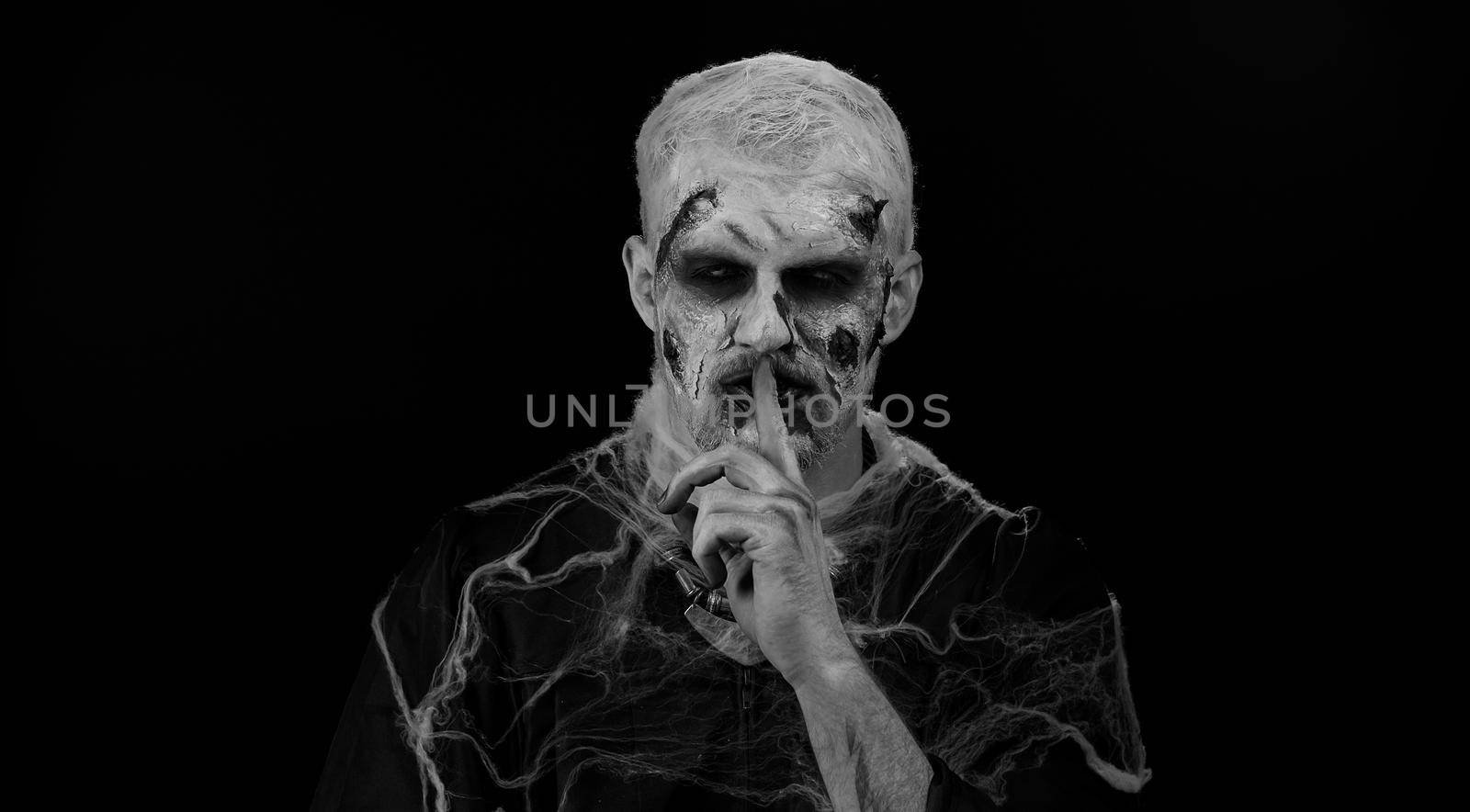 Shh be quiet. Sinister man with horrible scary Halloween zombie makeup in costume presses index finger to lips makes silence gesture sign do not tells secret. Dead guy with wounded bloody scars face