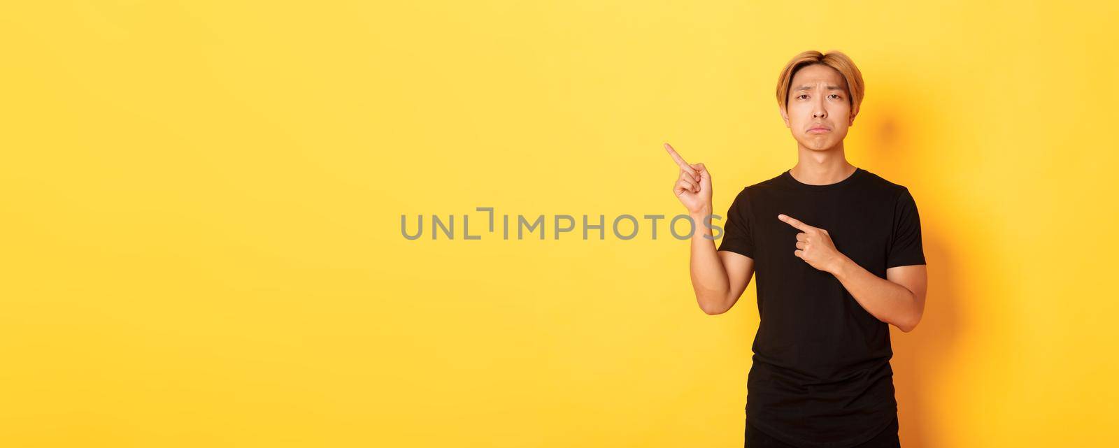 Portrait of misearble and gloomy asian guy in black t-shirt, pointing fingers upper left corner disappointed, yellow background by Benzoix