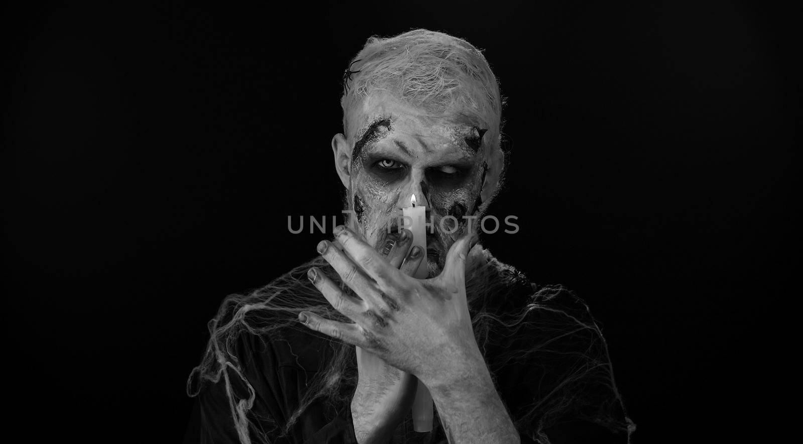 Zombie man with makeup with fake wounds scars and white contact lenses spells conjures over a candle, trying to scare in dark studio room. Sinister dead guy. Halloween, filming, staging concept
