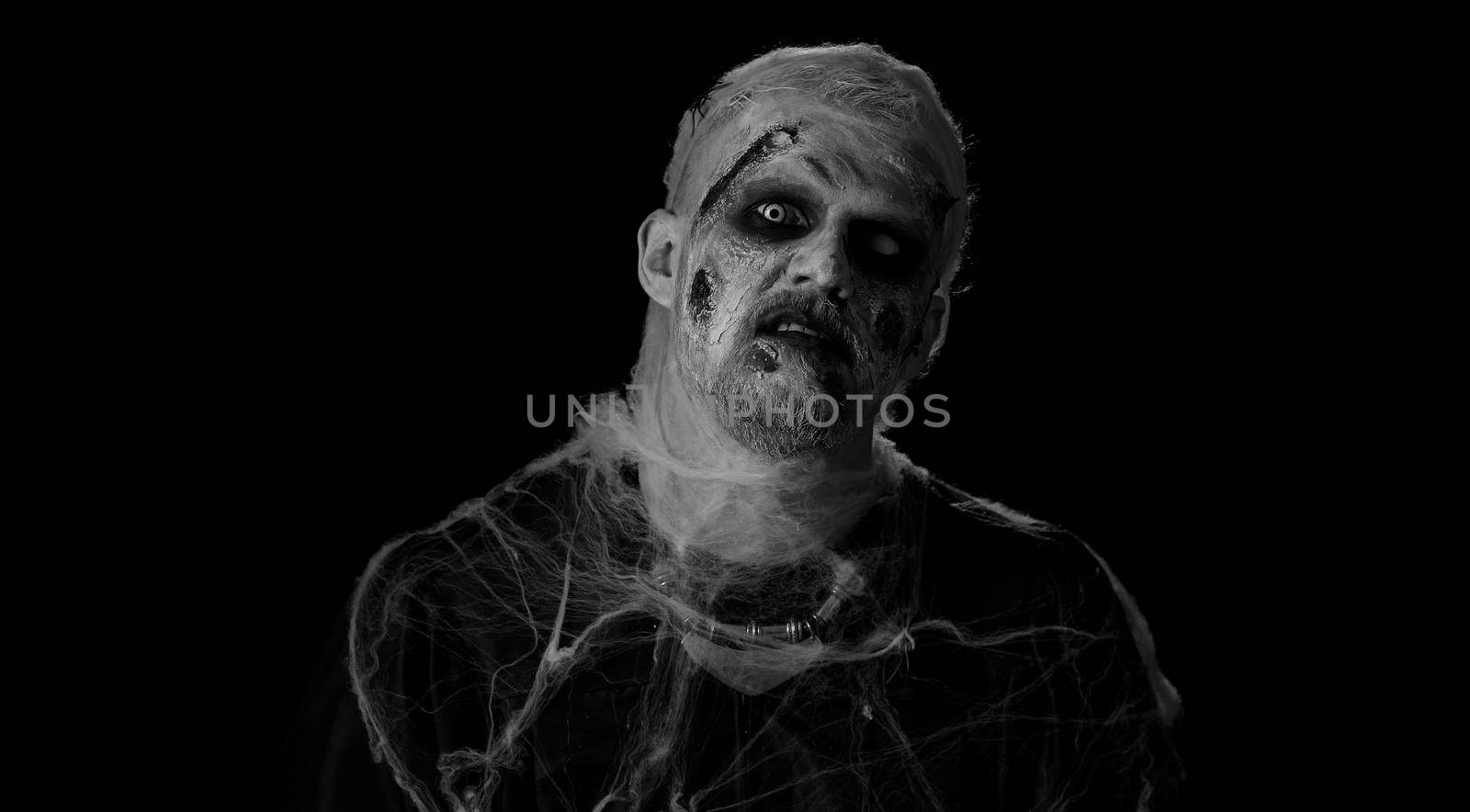 Sinister man with horrible scary Halloween zombie makeup in costume making faces, looking ominous at camera in police lights, trying to scare. Dead guy with wounded bloody scars face. Black background