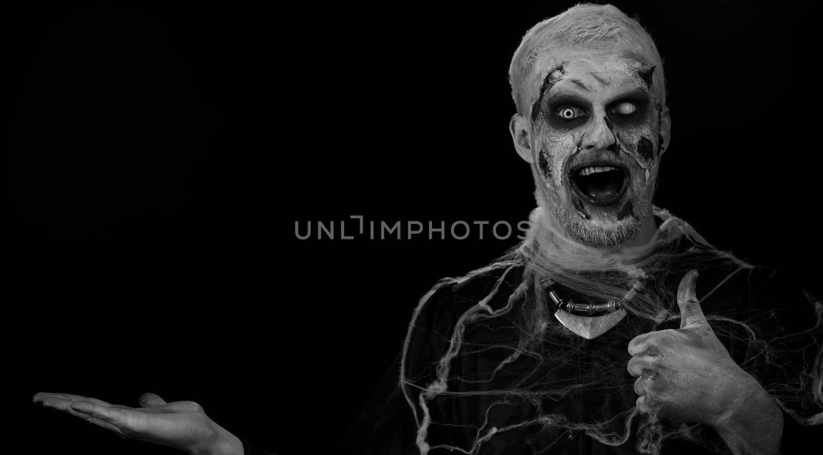 Zombie man with makeup with fake wounds scars showing thumbs up and pointing empty place, advertising area for commercial text logo, copy-space for goods promotion. Sinister Halloween ugly dead guy