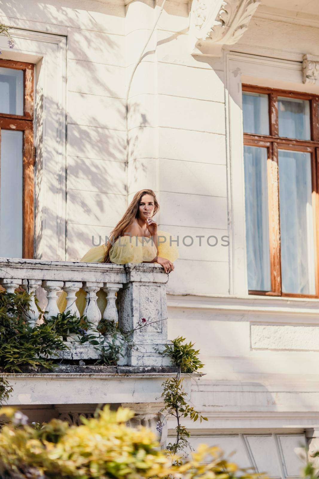 a beautiful smiling and kind woman in a gorgeous yellow dress stands on the balcony of an old vintage house.