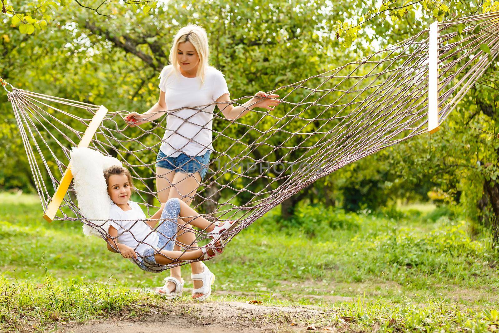 Happy mother and daughter relaxing together in a hammock at garden in sunny summer day. Family playing in hammock.