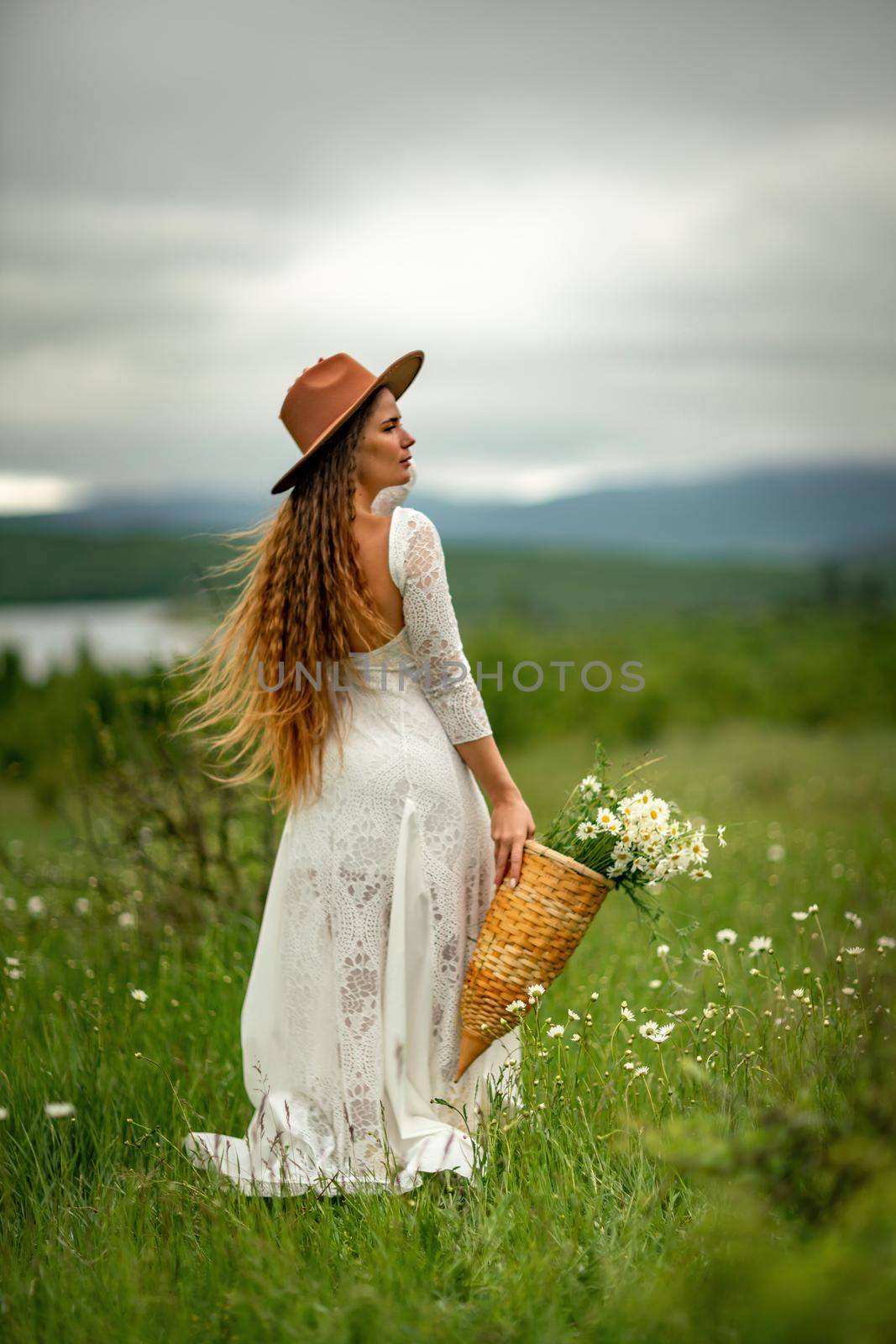 A middle-aged woman in a white dress and brown hat stands on a green field and holds a basket in her hands with a large bouquet of daisies. In the background there are mountains and a lake. by Matiunina