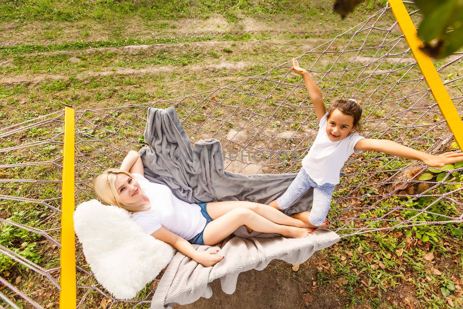 Beautiful young mother and daughter laying down and relaxing together on a hammock during a sunny summer day on holiday home garden. Family relaxing outdoors, healthy and wellness lifestyle.