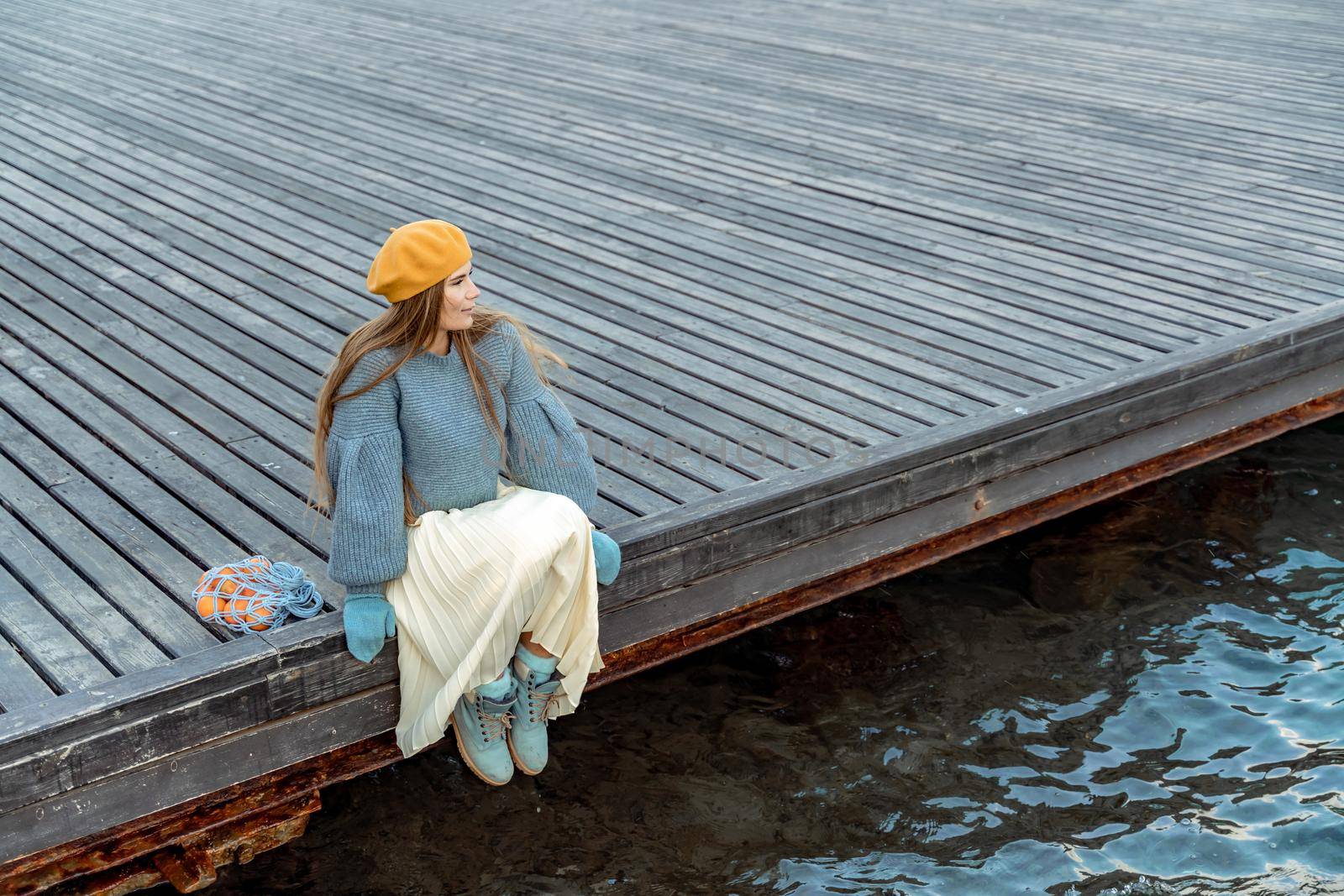 Outdoors fashion portrait of a beautiful middle aged woman walking on the beach. Marine background. Dressed in a stylish warm blue sweater, yellow skirt and beret