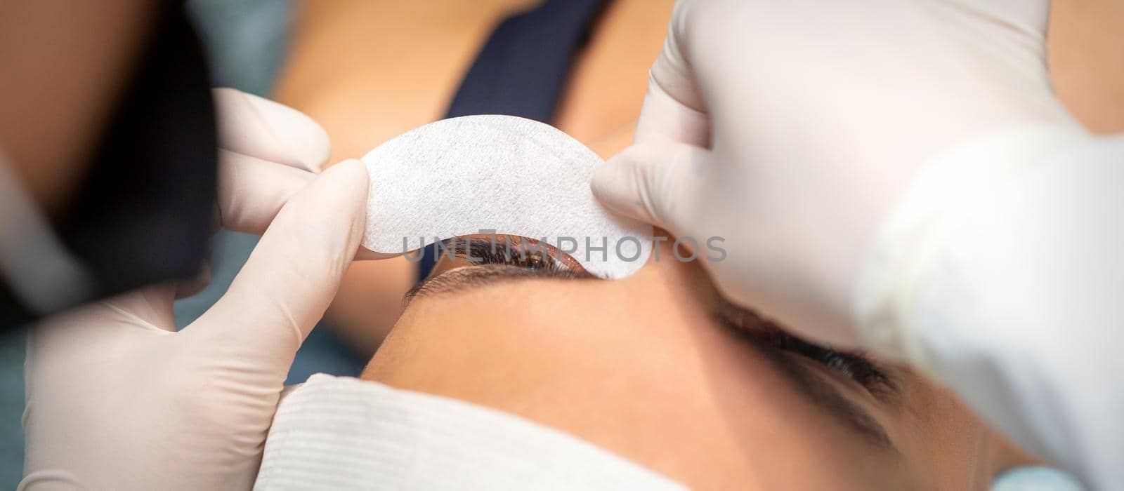 The beautician is gluing a patch under her eyes, before the procedure of eyelash extension. Artificial extended eyelashes