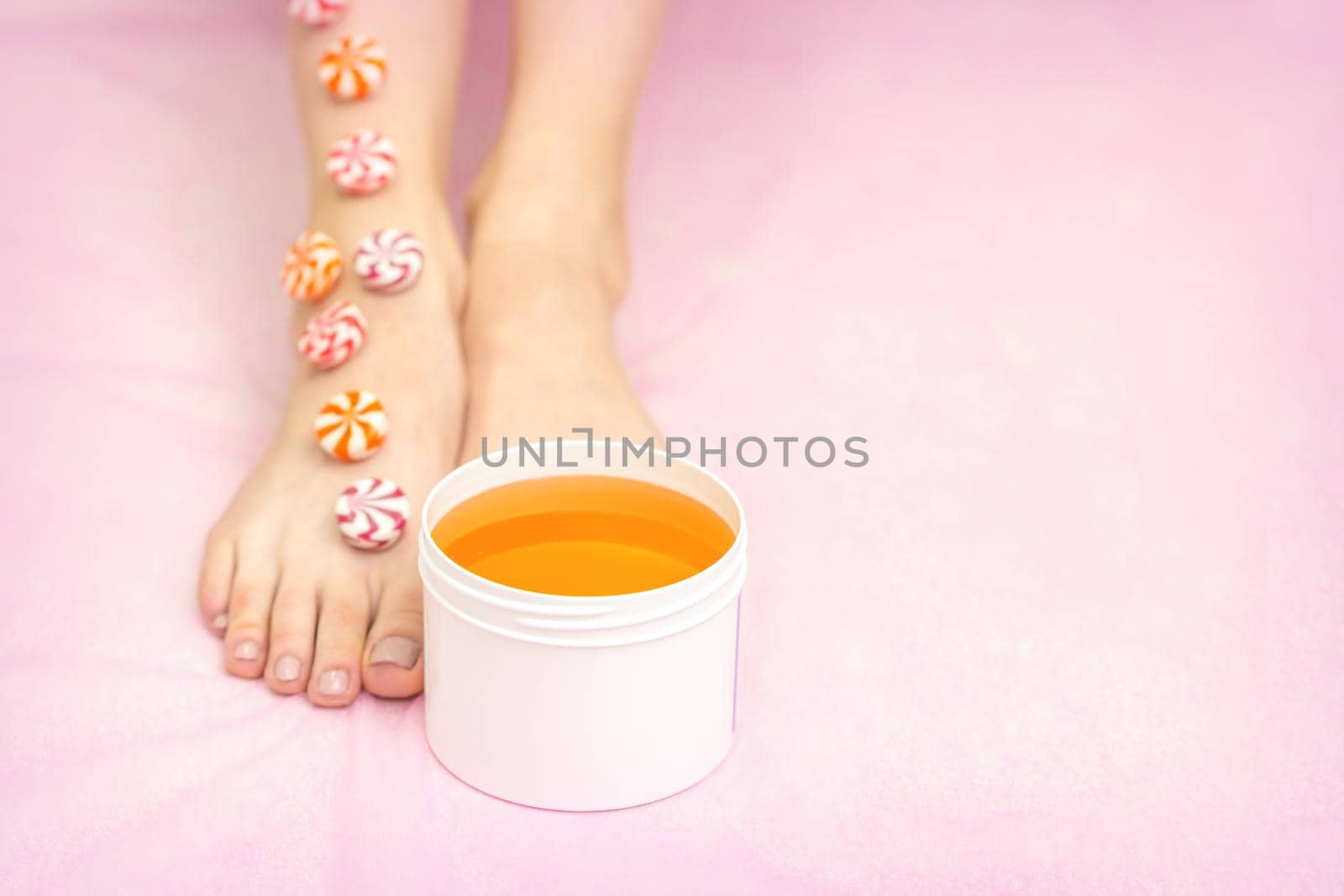 Round candies lying in a row on female feet with white jar with sugar paste on pink background with copy space, depilation concept