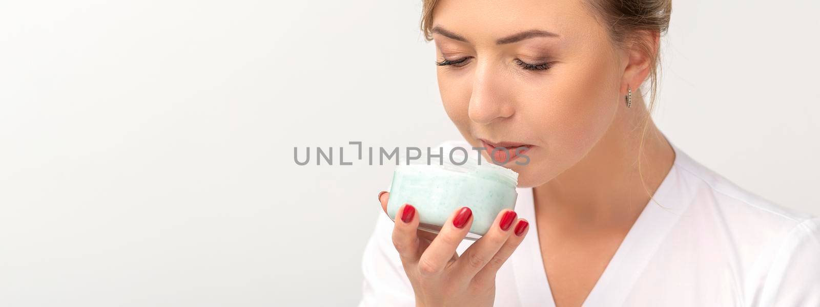 Portrait of young beautician sniffing moisturizing cream holding white jar on white background, copy space