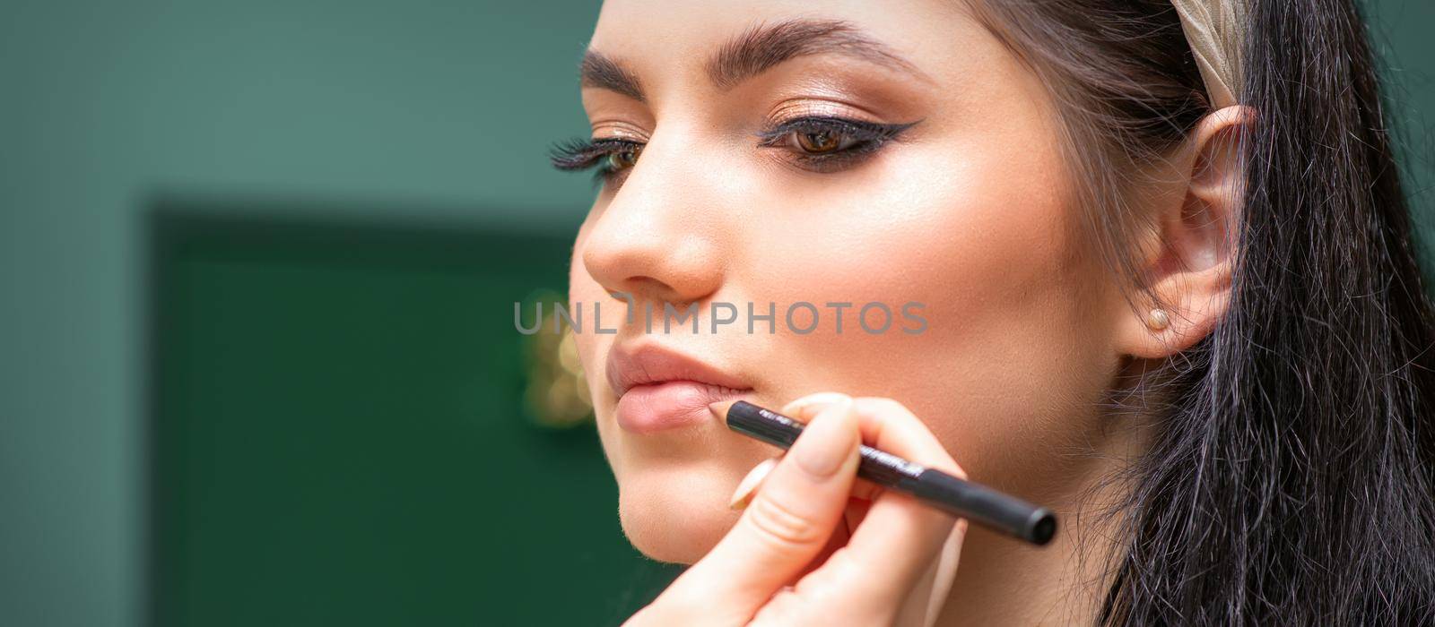 Makeup professional artist or cosmetologist is painting contour lips of a young woman with a pencil close up